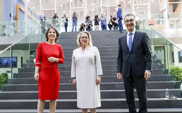 Drei Personen stehen lächelnd vor einer Treppe, während Fotografen im Hintergrund Bilder machen.