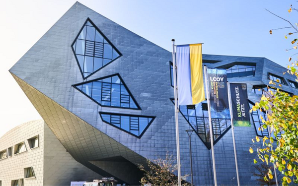 Ein futuristisches Gebäude mit geometrischen Fenstern und LCOY-Banner, flankiert von Fahnen, steht im Herbstlicht.