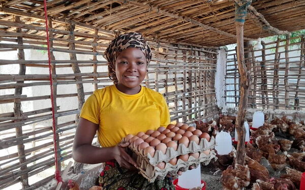 A person in a yellow shirt holds a tray of eggs, smiling inside a chicken coop filled with hens.