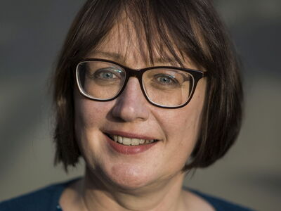 A woman with glasses smiles warmly, framed by soft lighting and wearing a blue top.