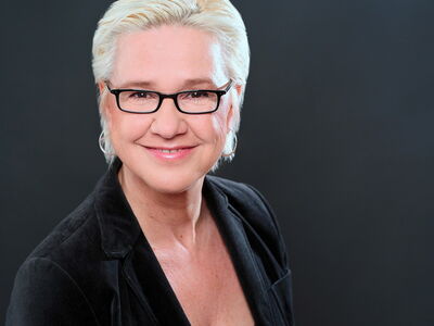 A person with short, blonde hair and glasses smiles warmly against a dark background, wearing a black blazer.