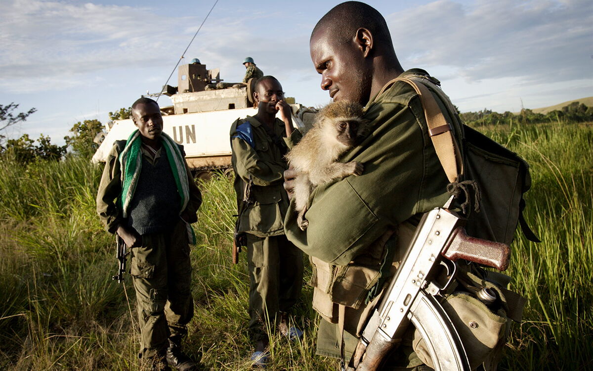 Ein Soldat hält ein kleines Äffchen im Arm, während zwei weitere Soldaten neben einem UN-Fahrzeug in einer grasbewachsenen Landschaft stehen.