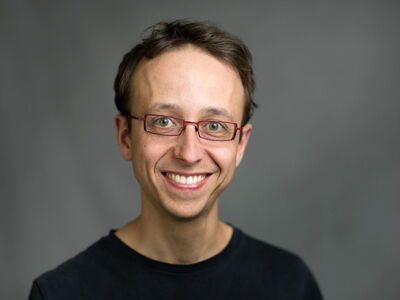 Ein Mann mit Brille lächelt freundlich in die Kamera vor einem neutralen grauen Hintergrund.