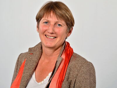 A woman in a brown jacket and orange scarf smiles warmly against a plain background.