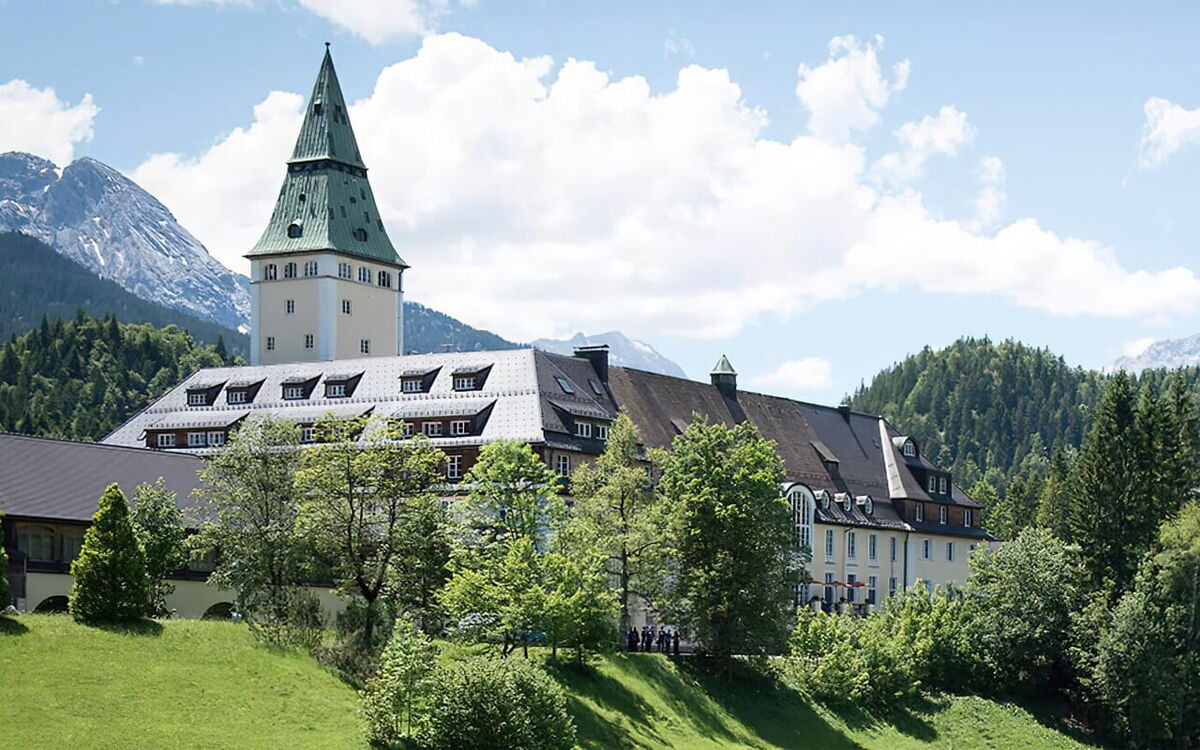 Ein malerisches Schloss in Elmau mit grünem Turm, umgeben von üppigen Wäldern und Bergen unter klarem Himmel.