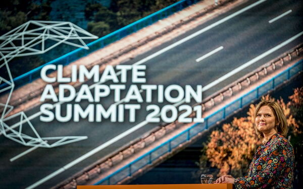 Eine Frau steht vor einem großen Bildschirm mit der Aufschrift "Climate Adaptation Summit 2021" und lächelt in die Kamera.
