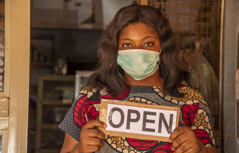 A woman wearing a face mask stands in a store entrance holding a sign that says "open".