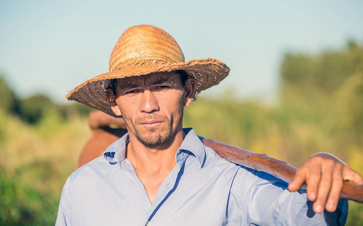 A man in a straw hat stands confidently in a sunny field, holding a tool over his shoulder against a blurred natural backdrop.