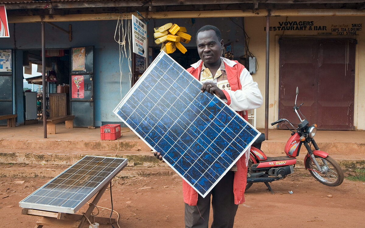 Ein Mann in roter Jacke präsentiert ein Solarpanel vor einem Geschäft, während ein Motorrad daneben steht.