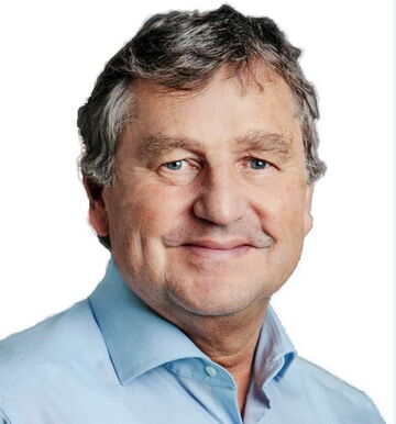 Ein lächelnder Mann in einem blauen Hemd schaut direkt in die Kamera vor einem weißen Hintergrund.