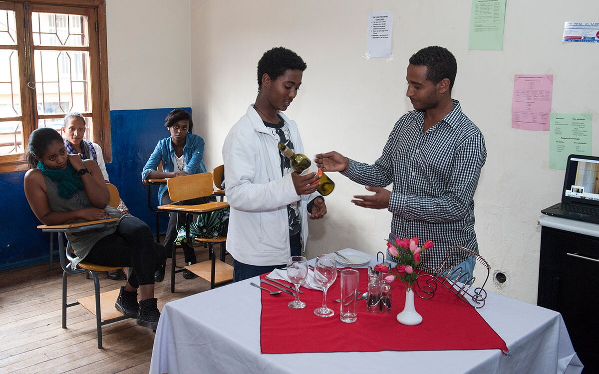 A young man practices wine pouring at a classroom table while students observe attentively, capturing a hands-on learning session.