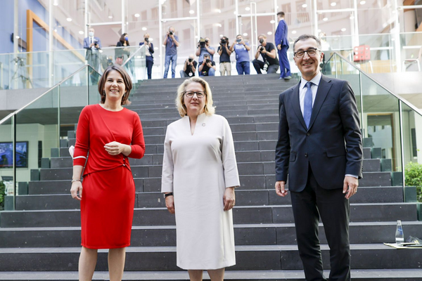 Three people stand smiling on a modern staircase with photographers capturing the moment, reflecting a formal event.