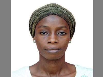 A woman wearing a patterned headscarf and earrings gazes directly at the camera against a plain background.