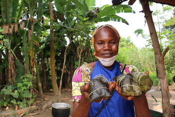 A woman holds wrapped food parcels, smiling in a lush garden setting with banana plants.