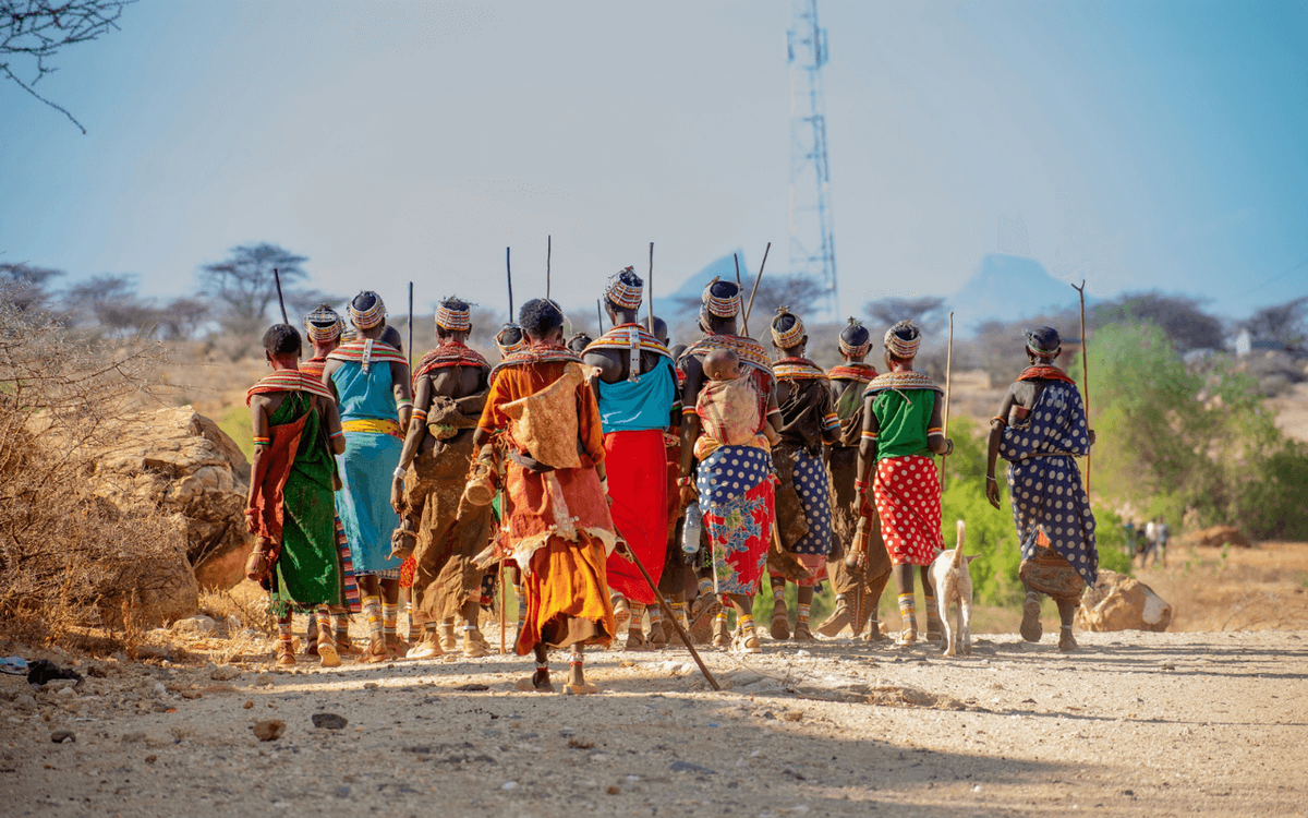 A group of traditionally dressed people, holding walking sticks, walk along a dusty path in a sunlit, rural landscape.
