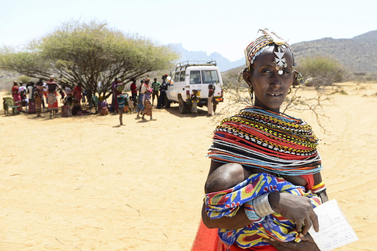 A woman in vibrant traditional attire holds a child, with a group gathered under a tree in a desert landscape.
