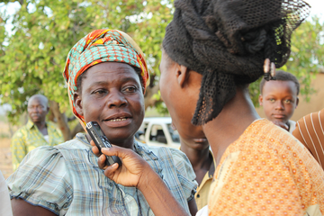 A woman in Ghana is being interviewed outdoors, holding a microphone, with onlookers in the background. | © Farm Radio International