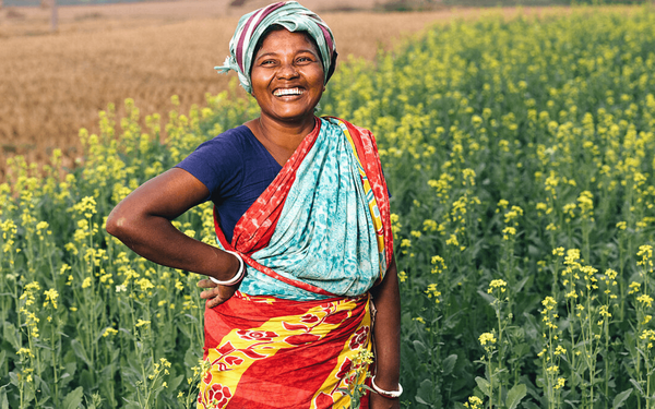 Eine lächelnde Frau in bunter Kleidung steht in einem blühenden Senffeld und genießt den sonnigen Tag.