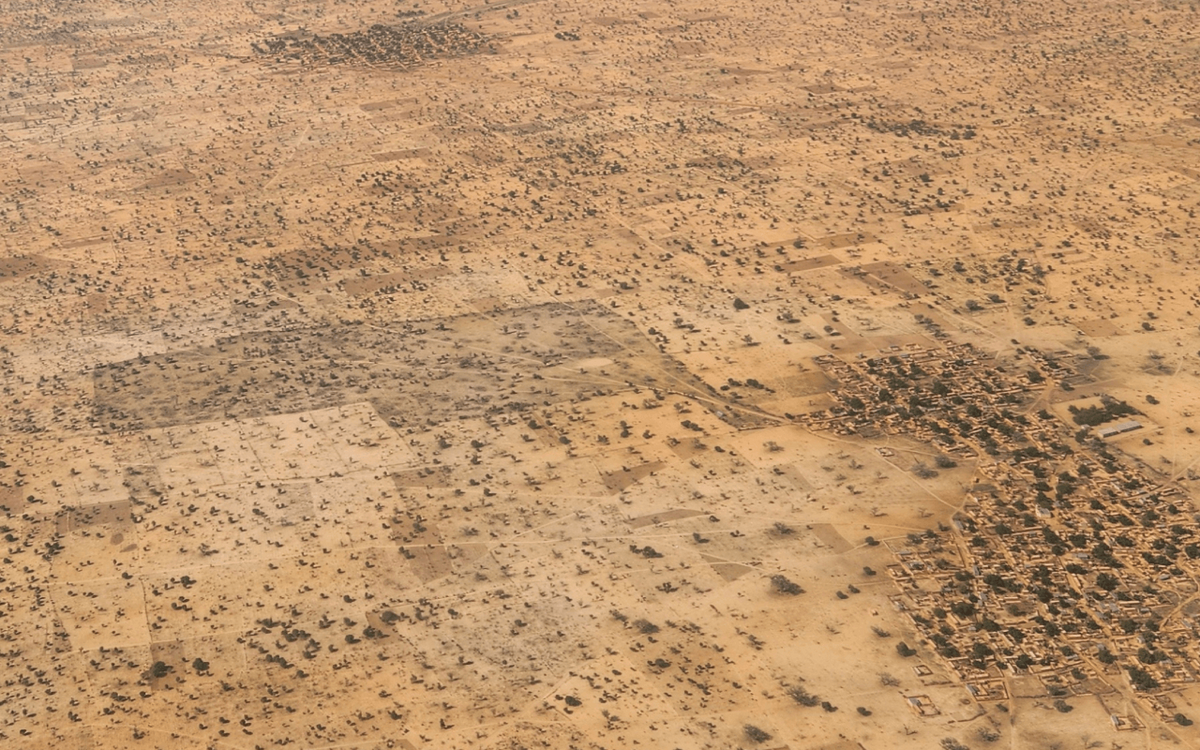 An aerial view shows a sparse desert landscape with scattered vegetation and a small clustered settlement in the distance.