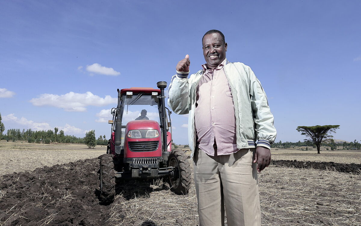 Ein lächelnder Mann steht vor einem roten Traktor auf einem frisch gepflügten Feld unter blauem Himmel.