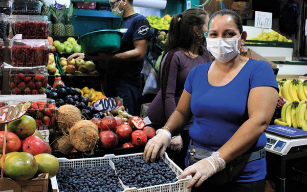A woman in a blue shirt and mask sells fresh blueberries at a vibrant rural market stand with various fruits.