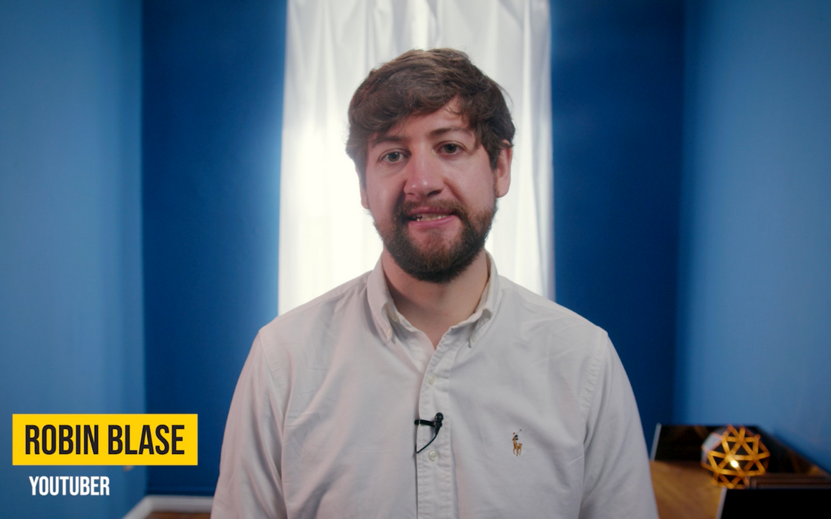 Ein Mann in einem weißen Hemd steht vor einem blauen Hintergrund und spricht in ein Mikrofon.