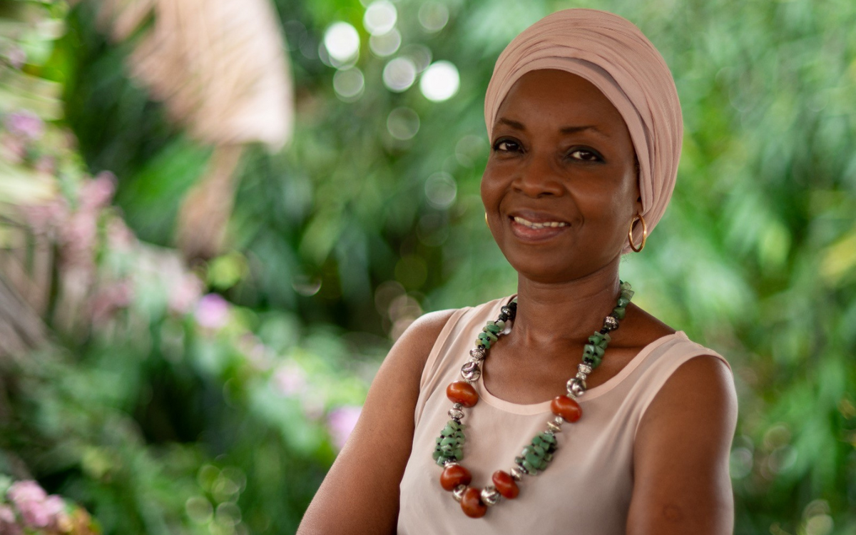 Eine lächelnde Frau mit Turban und bunter Halskette steht vor einem grünen, verschwommenen Hintergrund im Freien.