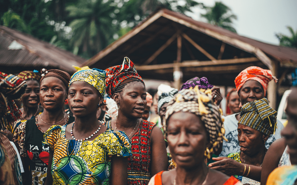 Eine Gruppe von Frauen in bunten traditionellen Kleidern und Kopfbedeckungen versammelt sich im Freien vor Hütten.