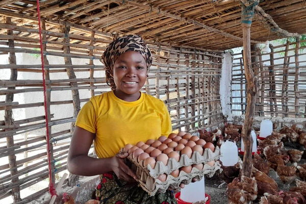 A person in a yellow shirt holds a tray of eggs, smiling inside a chicken coop filled with hens.